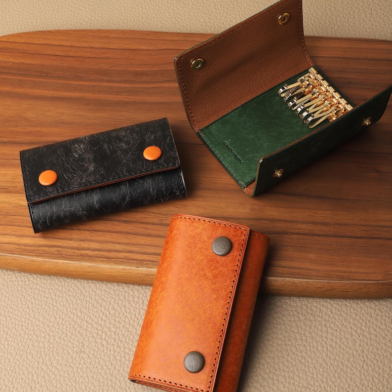 May include: Three leather key cases in black, brown, and orange are displayed on a wooden surface. The black case has an orange button closure. The brown case is open, revealing gold key hooks and a green interior. The orange case is closed with a dark button.