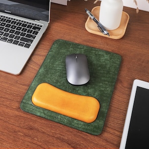 May include: A green leather mouse pad with an integrated orange wrist rest. A gray wireless mouse sits on the pad. A laptop and tablet are also visible on the wooden desk.