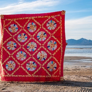 May include: A bright red quilt featuring a repeating floral pattern in white, yellow, and blue. The quilt is displayed against a beach scene with mountains and a blue sky in the background.