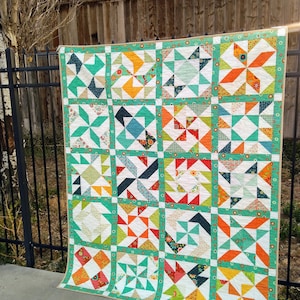 Puede incluir: Una colcha con un diseño de patchwork que presenta una variedad de telas coloridas. La colcha está hecha de cuadrados con diferentes patrones, incluyendo molinetes, estrellas y formas geométricas. La colcha es principalmente blanca con acentos de naranja, verde, rojo y azul.