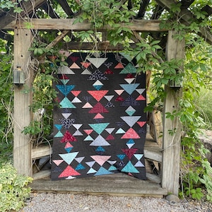 May include: A colorful quilt with a geometric pattern of triangles in shades of pink, blue, and green on a black background. The quilt is hanging on a wooden trellis covered in vines.