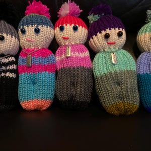 May include: Five knitted dolls with colorful striped bodies and pom-poms on their heads. Each doll has a silver chain with a tag that reads "Dream", "Trust", "Wonder", "Courage", and "Forgive".