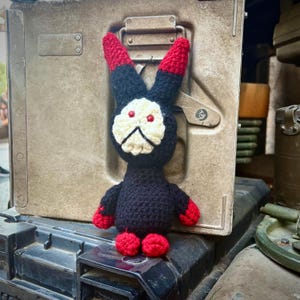 May include: A black and red crocheted bunny rabbit toy with white eyes and a sad expression. The toy is sitting on a metal surface.
