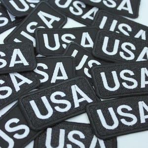 以下が含まれることがあります： 白い文字で「USA」と刺繍された黒い長方形のパッチの山。パッチはテクスチャ加工された表面と黒い縁取りがあります。白い背景に散らばっています。