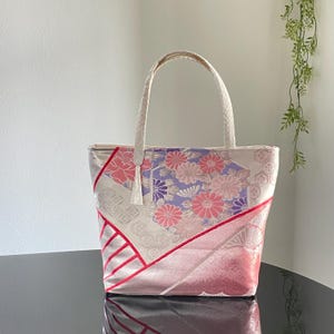 以下が含まれることがあります： クリーム色のハンドルとタッセルが付いた、花柄のトートバッグ。 バッグは、クリーム色の背景にピンクと紫の花のパターンがあり、赤いアクセントが付いています。 バッグは、テクスチャード加工された生地で作られています。