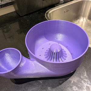 Op de afbeelding: Een lavendelkleurige 3D-geprinte citruspers met een grote ronde kom, een centrale perskegel en een kleinere tuit voor het uitschenken van sap. Het apparaat heeft een gladde, matte afwerking en een functioneel ontwerp voor het extraheren van sap.