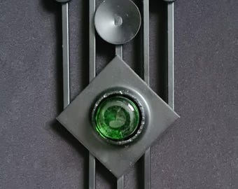 May include: A black metal wall sconce with a geometric design. It features a central diamond shape with a green glass insert, surrounded by vertical lines and circular accents. The sconce has a small hook for hanging and a candle holder.