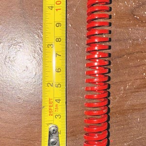 Pode incluir: Uma mola helicoidal vermelha brilhante, com cerca de 10 cm de comprimento, é colocada ao lado de uma fita métrica amarela. A fita mostra medidas em pés e polegadas, com o comprimento da mola claramente visível.