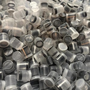 May include: A pile of small, clear plastic containers with black and silver lids. The containers are cylindrical and appear to be stacked together. The lids have a circular design. The overall color scheme is clear, black, and silver.