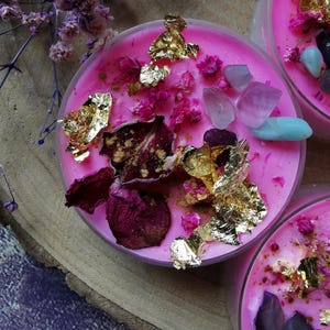 Könnte beinhalten: Nahaufnahme von drei rosa Kerzen mit dekorativen Elementen. Jede Kerze ist mit getrockneten Blumen, Blattgold und Edelsteinen verziert. Die Kerzen stehen auf einer Holzoberfläche, was eine natürliche und elegante Ästhetik erzeugt.