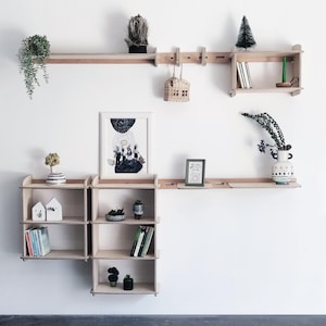 Op de afbeelding: Een set houten wandplanken met verschillende planten, een ingelijst drukwerk en boeken. De planken zijn gemaakt van natuurlijk hout en hebben een eenvoudig, modern ontwerp.