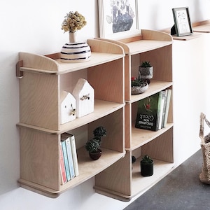 May include: Wooden modular shelves with a natural finish, mounted on a white wall. The shelves display decorative items like small houses, potted plants, books, and a vase with dried flowers. The design is minimalist and modern, suitable for home decor.