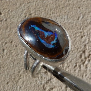 Peut inclure: Une bague en argent avec une pierre ovale en opale brun foncé sertie au centre. L'opale présente un éclat irisé bleu et blanc.