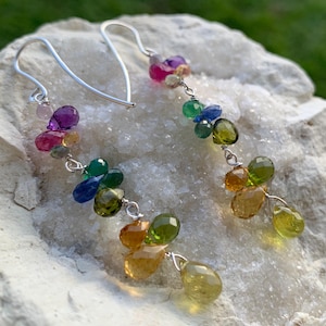 May include: A pair of dangle earrings featuring a cascade of colorful gemstones. The earrings have a silver wire hook and are adorned with a variety of faceted stones in shades of pink, purple, green, blue, and yellow. The gemstones are strung together with silver wire.