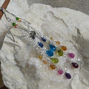 May include: A pair of silver dangle earrings with a rainbow of gemstone drops. The earrings feature a variety of colors, including blue, green, yellow, orange, red, and purple.