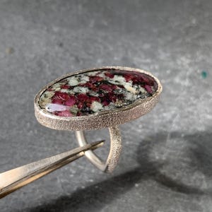 Op de afbeelding: Een zilveren ring met een grote ovale edelsteen. De edelsteen is een mix van witte, zwarte en rode kleuren.