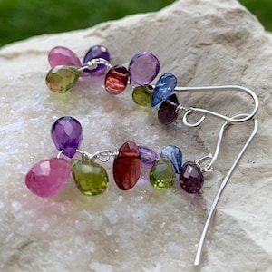 May include: Dangling earrings with a cluster of colorful gemstones. The earrings feature a variety of faceted stones in shades of pink, purple, green, red, and blue, strung together with silver wire and finished with silver ear hooks.