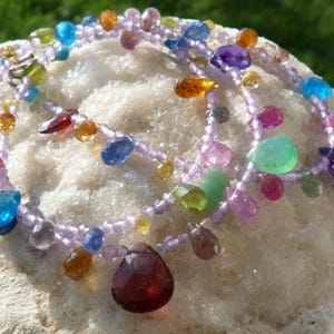 May include: A multi-strand necklace with a variety of colorful gemstones, including amethyst, sapphire, ruby, and citrine. The necklace is made with a light purple bead string and features a large, teardrop-shaped garnet pendant.