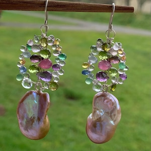 May include: A pair of dangle earrings featuring large, iridescent, teardrop-shaped pearls. The earrings have a cluster of colorful gemstones in shades of green, pink, purple, and clear, suspended from silver hooks. The background is a blurred green.