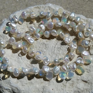 Peut inclure: Un collier composé de perles d'eau douce blanches, irrégulières et lustrées, entremêlées de petites opales translucides et irisées. Le bijou présente un fermoir à crochet en métal doré et est disposé en cercle avec une surface réfléchissante.
