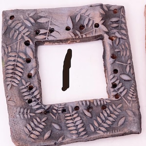 May include: A square, ceramic frame with a gray-brown finish. The frame is decorated with a leaf pattern and small holes. The center of the frame is white with a black, vertical line.