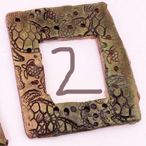 May include: A rectangular, ceramic frame with a square opening. The frame is primarily olive green with brown accents and features a turtle design. The number "2" is drawn in the center of the frame. The frame has several small holes.