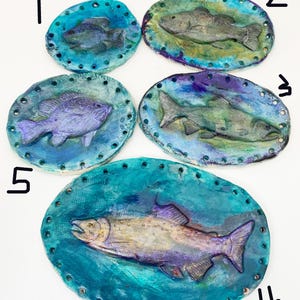 May include: Five handmade ceramic fish plaques. Each plaque features a different fish design in shades of blue, purple, and gray. The plaques are oval-shaped with a textured border and are suitable for wall decor.