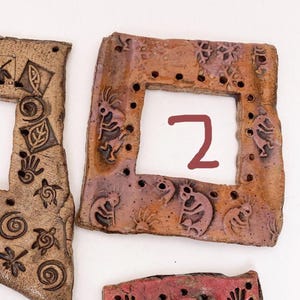 May include: A square, terracotta-colored ceramic frame featuring embossed figures of flute players and snowflake patterns. The frame has a series of small, evenly spaced holes along the inner border and the number 2 written in red.
