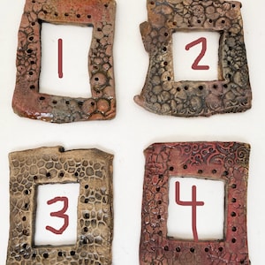 May include: Four handmade ceramic tiles with numbers 1, 2, 3, and 4. Each tile has a rectangular frame with a textured, earthy design in shades of brown, red, and tan. The numbers are hand-drawn in a dark red color.