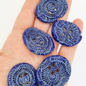 May include: Five oval-shaped ceramic buttons with a glossy deep blue glaze. Each button features a textured, radiating spiral pattern and four central holes for attachment. The surface has a slightly irregular, handcrafted appearance.