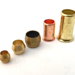 May include: An assortment of brass and copper fittings, including compression fittings and a ferrule, are arranged on a white surface. The fittings vary in size and shape, with a mix of cylindrical and flared designs. The copper fitting has a reddish-brown hue.