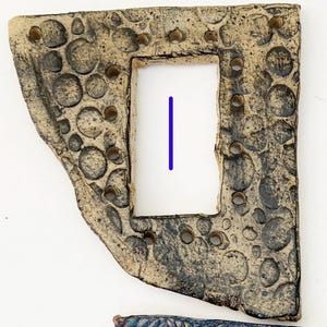 May include: A ceramic tile with a rectangular opening in the center. The tile is decorated with a pattern of small circles and has a rough, textured surface.