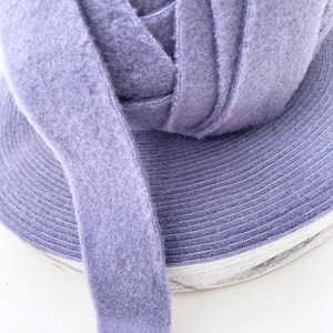 May include: Close-up of a roll of lavender-colored fabric ribbon. The ribbon is made of a soft, fuzzy material and is tightly wound. The edges of the ribbon are stitched with a white thread. The background is white.