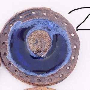 May include: A round, brown ceramic dish with a blue and white glaze. The dish has a ring of holes around the edge and a smaller circle in the center.