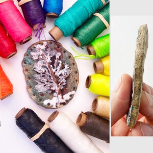 May include: A handmade, ceramic, leaf-shaped pendant with a textured surface, held by a hand. The pendant is surrounded by colorful spools of thread in various colors, including red, pink, green, and yellow. The pendant is approximately 3 inches long.