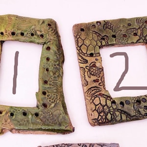 May include: Two square ceramic frames with a textured, mottled green and brown finish. The frames have decorative turtle and geometric patterns. The number "1" and "2" are written inside the frames.