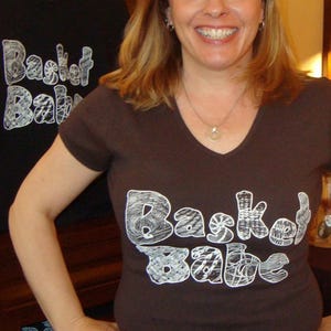 May include: A woman wearing a brown t-shirt with the text "Basket Babe" printed in white. The text is stylized with a basket weave pattern.