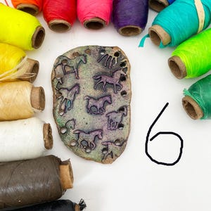 May include: A collection of colorful spools of thread surrounds a textured, irregular-shaped pendant. The pendant features embossed horse designs. The threads are in various colors, including red, pink, yellow, and green.