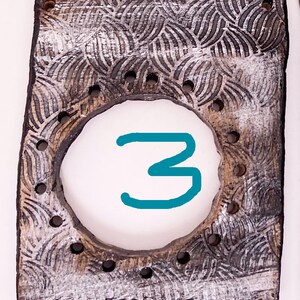 May include: A wooden tile with a white and brown patterned design. The tile has a circular cutout with the number "3" written in teal blue.
