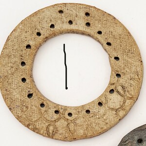 May include: A circular, tan-colored, textured object with a series of small, evenly spaced holes punched around its perimeter. The surface features a subtle, repeating embossed pattern. A thin, black vertical line is drawn inside the center opening.
