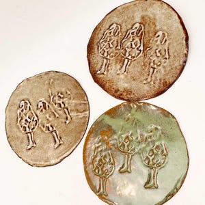 May include: Three ceramic plates in shades of brown and green. Each plate features an embossed design of three stylized mushrooms. The plates have irregular, organic shapes and varying textures, adding to their handmade aesthetic.
