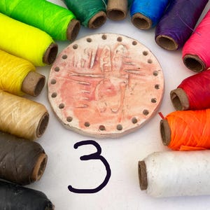 May include: A collection of colorful spools of thread arranged around a circular, reddish-brown ceramic piece with carved designs and small holes. The number "3" is written in black ink below the ceramic piece.