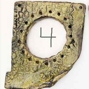 May include: A rectangular ceramic piece with a large circular cutout in the center and the number 4 written in the middle. The surface features a textured, crackled, light green and brown glaze with a series of small holes along the inner circle.