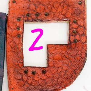 May include: A terracotta-colored ceramic tile with a textured surface and a cutout in the shape of the letter "Z". The tile has several small holes around the edges and a brown glaze.