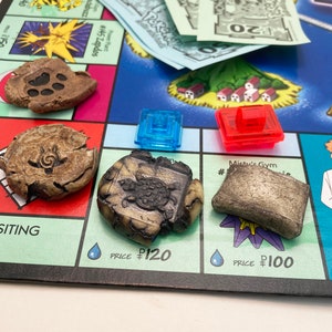 May include: A close-up of a game board with four tokens. The tokens are made of a brown, textured material and feature different designs. The board is blue and green and features the text "Misty's Gym" and "Price $100".