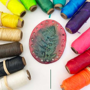 May include: An assortment of colorful spools of thread surrounds a red and green leather piece with a fern design. The thread colors include yellow, green, blue, purple, pink, red, orange, black, and white.