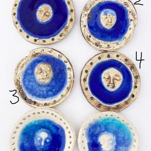 May include: Six ceramic round plates with a blue glaze and a white face design in the center. The plates have a white rim with small holes around the edge.
