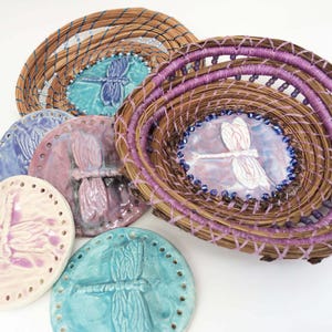 May include: Five ceramic dragonfly ornaments with a woven basket. The ornaments are in shades of pink, blue, and white. The basket is woven with brown and purple reeds.