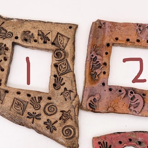 May include: Two handcrafted ceramic frames with square openings, numbered 1 and 2. The frames are textured with a brown and reddish-brown color scheme. Each frame is adorned with carved designs of insects, leaves, and other symbols.