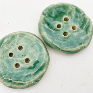 May include: Two round, handmade ceramic buttons with a glossy, variegated seafoam green glaze. Each button features an irregular, organic shape and four central holes for attachment, showcasing a smooth, reflective surface texture.